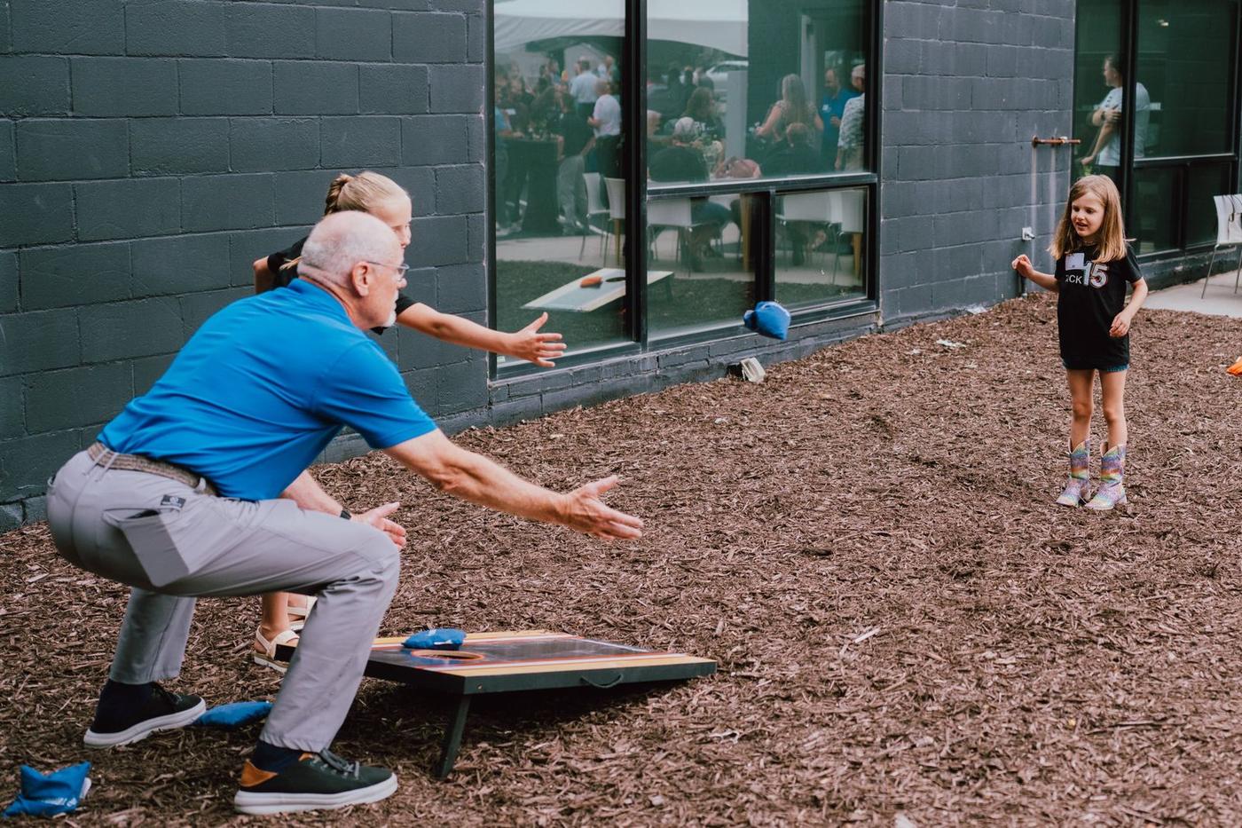 A guest and two kids playing cornhole beside the TACK building