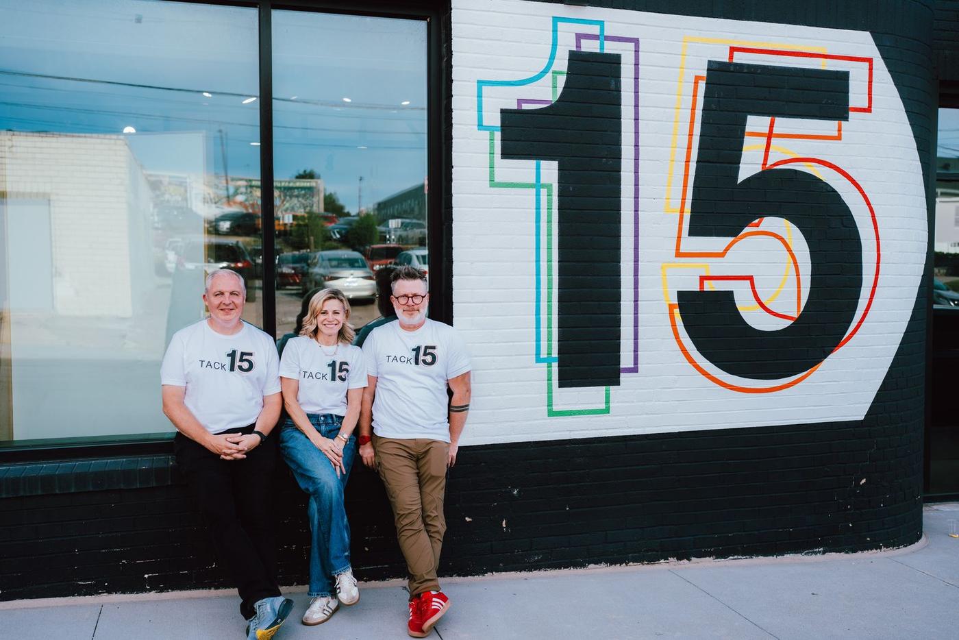Three members of TACK in anniversary shirts in front of a large painted 15 mural